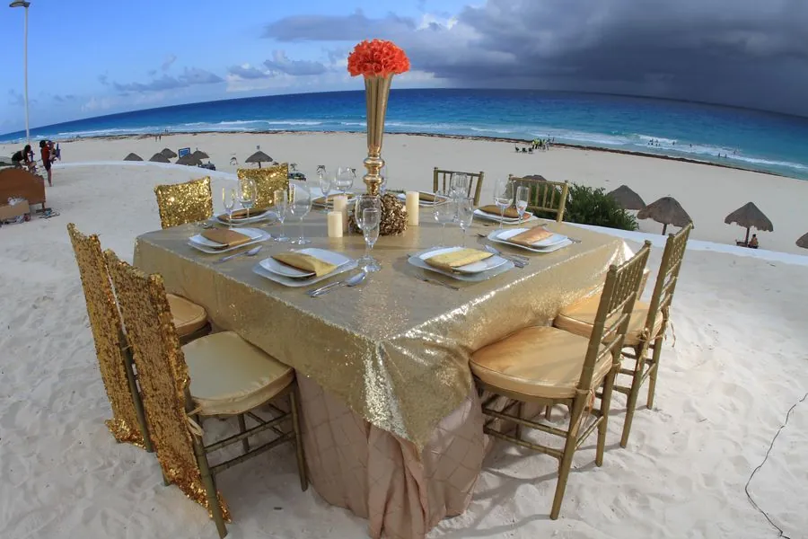 Mesa cuadrada en la playa con mantel dorado y sillas Tiffany doradas