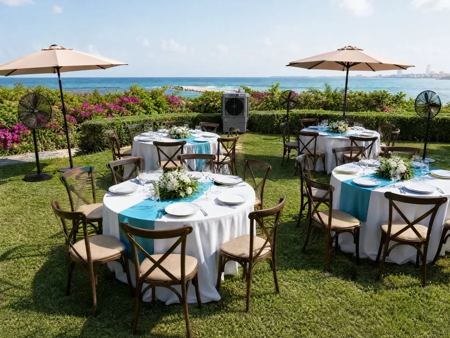 Mesas redondas con mantel blanco y sillas crossback en un jardín frente al mar, con sombrillas, ventiladores de pedestal y un portacooler