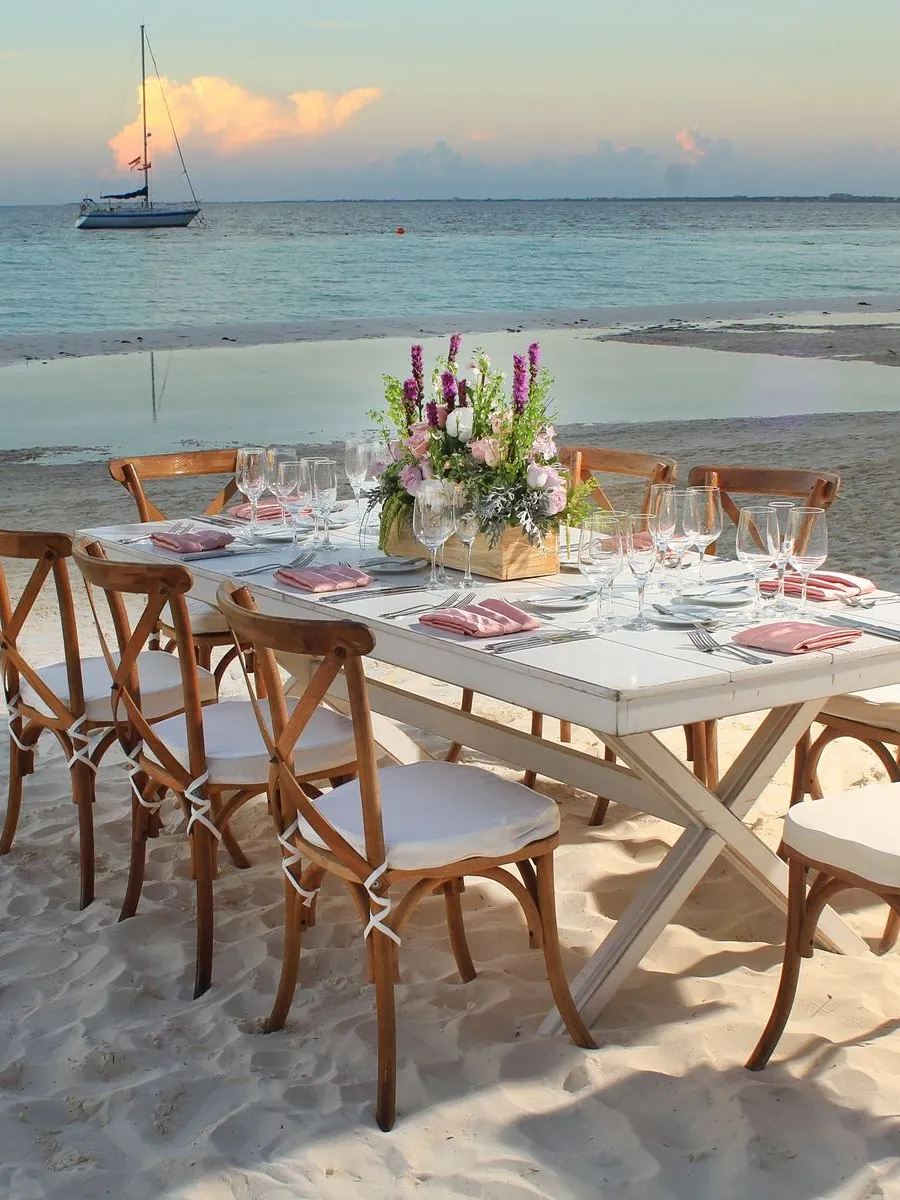 Mesa blanca en la playa al atardecer con sillas crossback de madera, copas y centro de flores